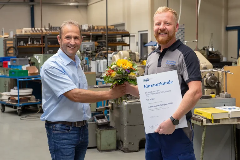 René Liebermann überreicht Tim Grübel einen Strauß Blumen zu seinem Jubiläum, beide lächeln in die Kamera