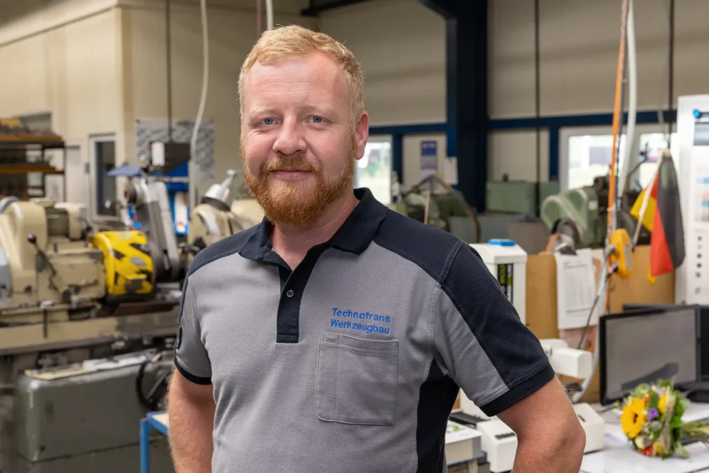 Portaitfoto Tim Grübel: lächelnder Herr mit blond-roten Haaren und Vollbart im Technotrans-Poloshirt, im Hintergrund Maschinen